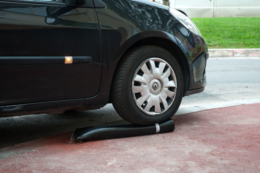 A black vehicle with a scuffed wheel runs over a bollard that bends 90 degrees