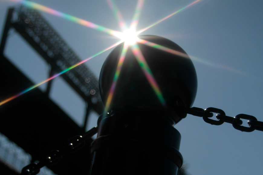 A bollard, shot from the ground, glints with sunlight and is framed by a blue sky and baseball diamond lighting