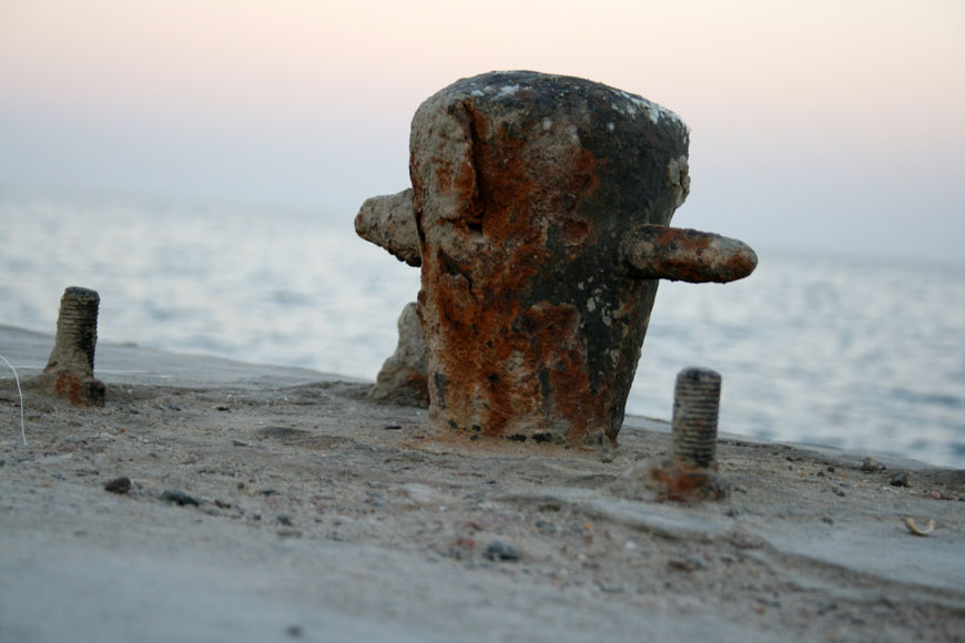 Rusty bollard by the ocean, narrower on the bottom and wider on top, with two short protruding pegs